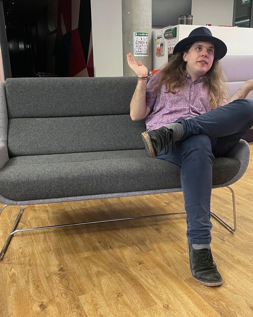 Zuri mit Hut auf einem grauen Sofa in einem Büro, gestikuliert beim Erklären
